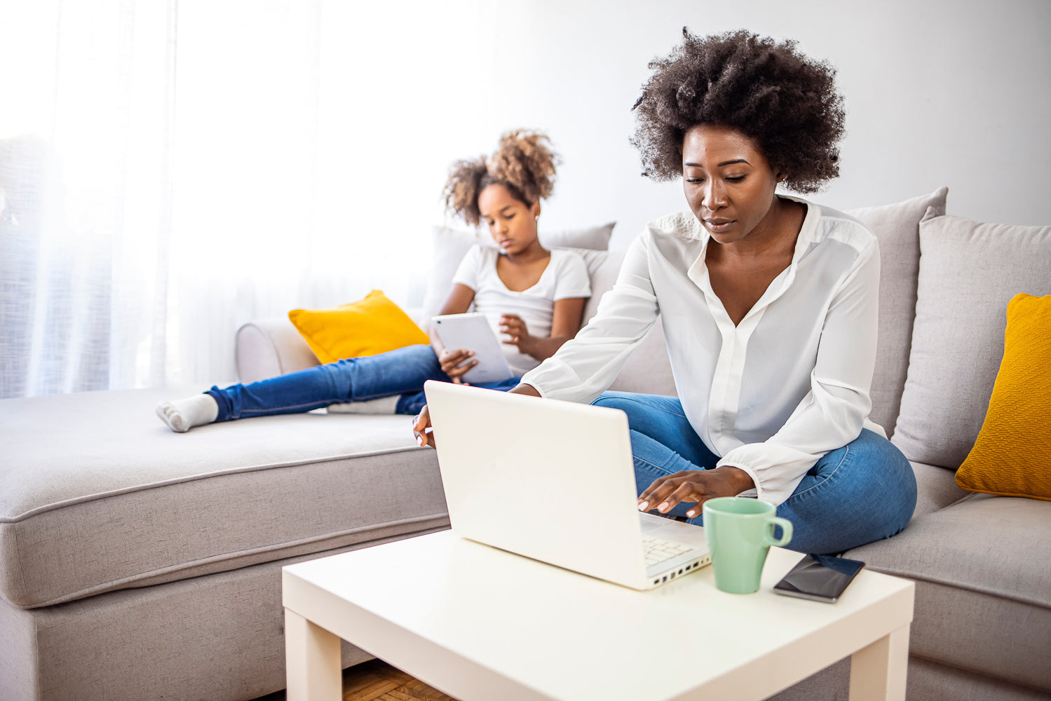 Black mother sitting on couch working on laptop, daughter sitting on couch behind her on tablet